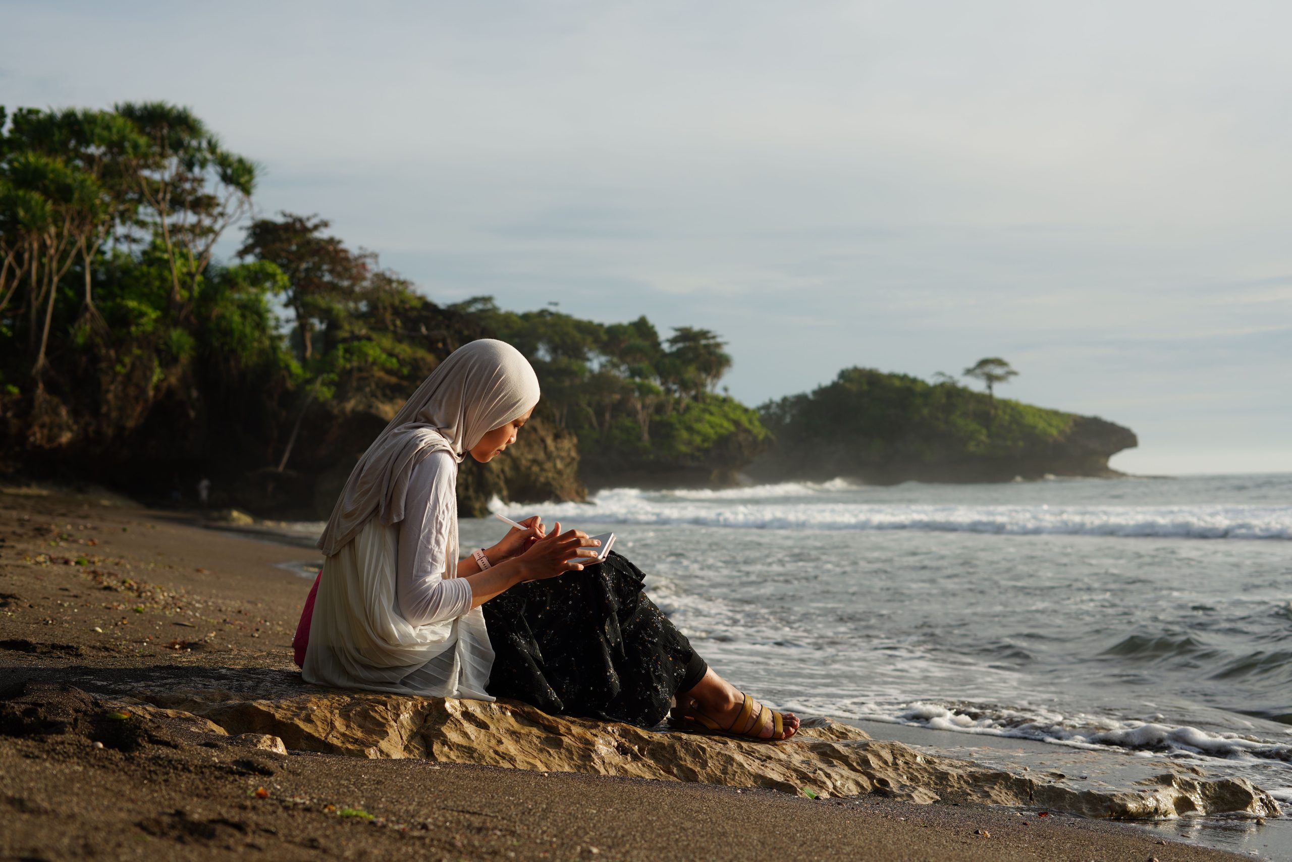 A woman sitting on a rock at the shoreline, writing in a notebook as the waves come in.